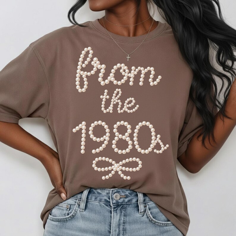 May include: A brown t-shirt with the text "From the 1980s" in a pearl-like font. The shirt has short sleeves and a round neckline. The text is arranged in a decorative style with a bow at the bottom.