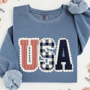 May include: A blue crew neck sweatshirt with the word "USA" appliqued across the front. The "U" has red and white stripes, the "S" is a blue and white gingham pattern, and the "A" is navy blue with white polka dots. The letters are outlined with white lace.