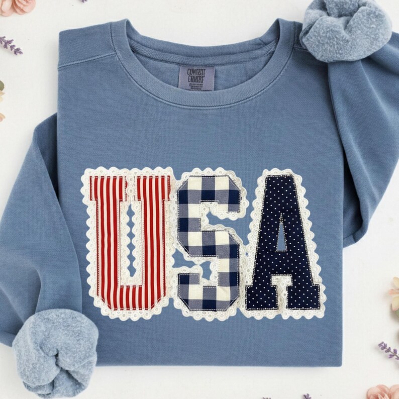 May include: A blue crew neck sweatshirt with the word "USA" appliqued across the front. The "U" has red and white stripes, the "S" is a blue and white gingham pattern, and the "A" is navy blue with white polka dots. The letters are outlined with white lace.