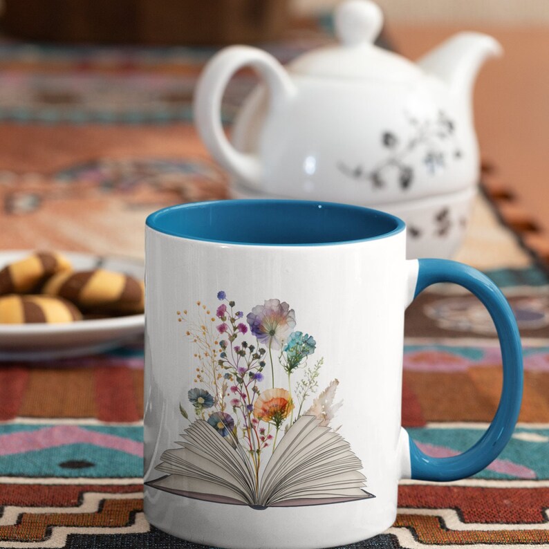 Pu&ograve; includere: Una tazza in ceramica bianca con un bordo e un manico blu. La tazza presenta un motivo floreale colorato con un libro aperto sotto i fiori. Il libro &egrave; aperto e ha pagine con delle righe.