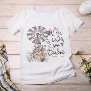 May include: White t-shirt with a watercolor design of a windmill, flowers, and butterflies. The text "Life is better in a small town" is printed on the shirt.