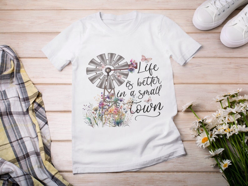 May include: White t-shirt with a watercolor design of a windmill, flowers, and butterflies. The text "Life is better in a small town" is printed on the shirt.
