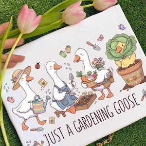 May include: White rectangular decorative pillow with a whimsical illustration of gardening geese. The geese are depicted with gardening tools, vegetables, and flowers. The text "JUST A GARDENING GOOSE" is printed on the pillow.