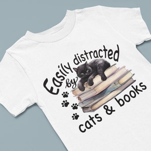 May include: White t-shirt with a black cat sleeping on a stack of books. The text reads "Easily distracted by cats & books".
