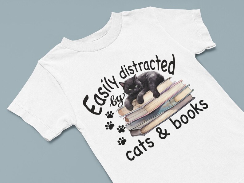May include: White t-shirt with a black cat sleeping on a stack of books. The text reads "Easily distracted by cats & books".