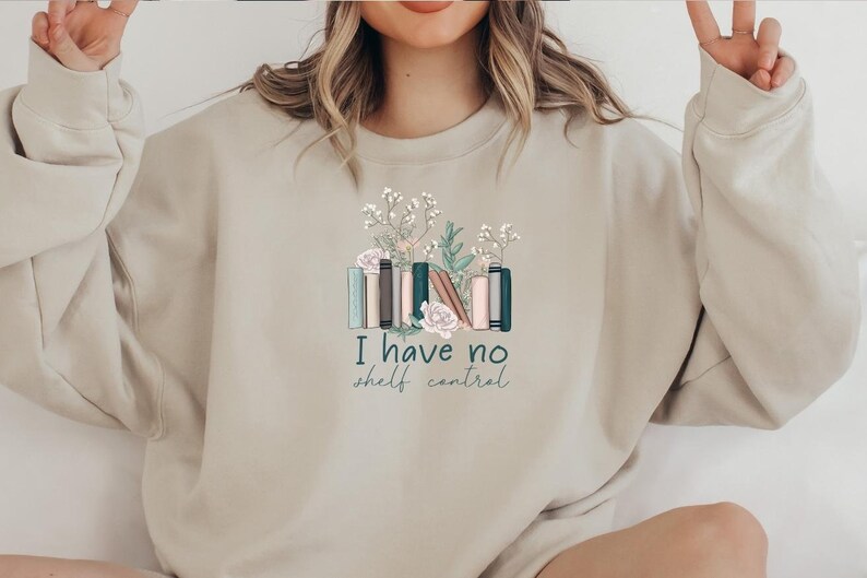May include: A beige sweatshirt with a graphic of a stack of books with flowers and the text "I have no shelf control".
