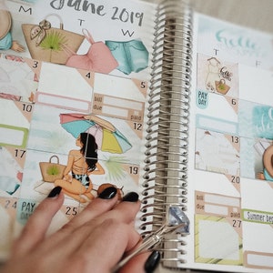 K&ouml;nnte beinhalten: Ein Planer mit einem Kalender f&uuml;r Juni 2019. Der Planer ist mit Aufklebern verziert, die Strandmotive zeigen, darunter eine Frau im Badeanzug, ein Sonnenschirm und ein Strohhut. Der Planer hat eine silberne Spiralbindung.