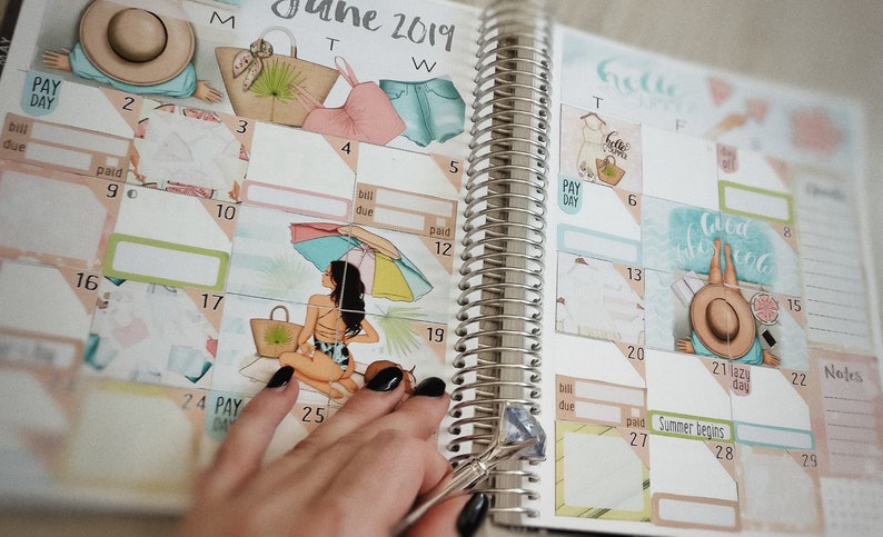 K&ouml;nnte beinhalten: Ein Planer mit einem Kalender f&uuml;r Juni 2019. Der Planer ist mit Aufklebern verziert, die Strandmotive zeigen, darunter eine Frau im Badeanzug, ein Sonnenschirm und ein Strohhut. Der Planer hat eine silberne Spiralbindung.