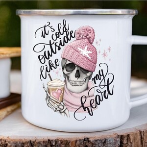 May include: White enamel mug with a silver rim featuring a skull wearing a pink knit hat and scarf. The skull is holding a coffee cup with a heart. Text on the mug reads "It's cold outside like my heart."