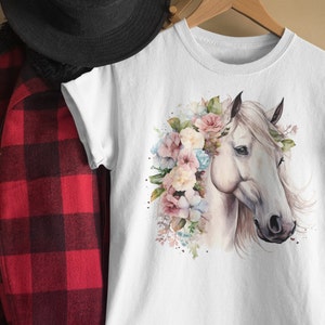 May include: White t-shirt with a watercolor illustration of a white horse with a floral crown. The flowers are pink, blue, and white.