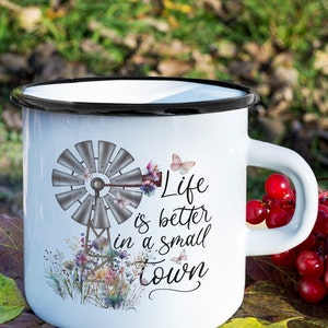 May include: White enamel mug with a black rim and handle. The mug features a watercolor illustration of a windmill with flowers and butterflies. The text "Life is better in a small town" is printed on the mug.