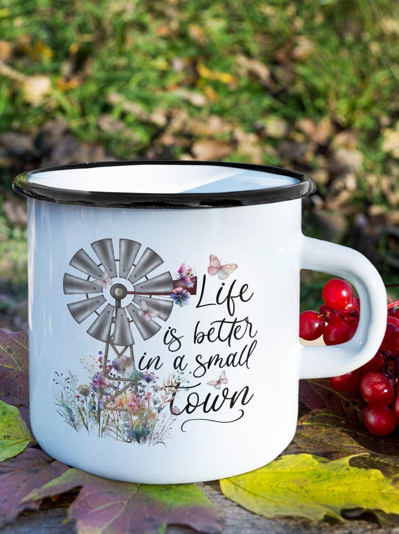 May include: White enamel mug with a black rim and handle. The mug features a watercolor illustration of a windmill with flowers and butterflies. The text "Life is better in a small town" is printed on the mug.