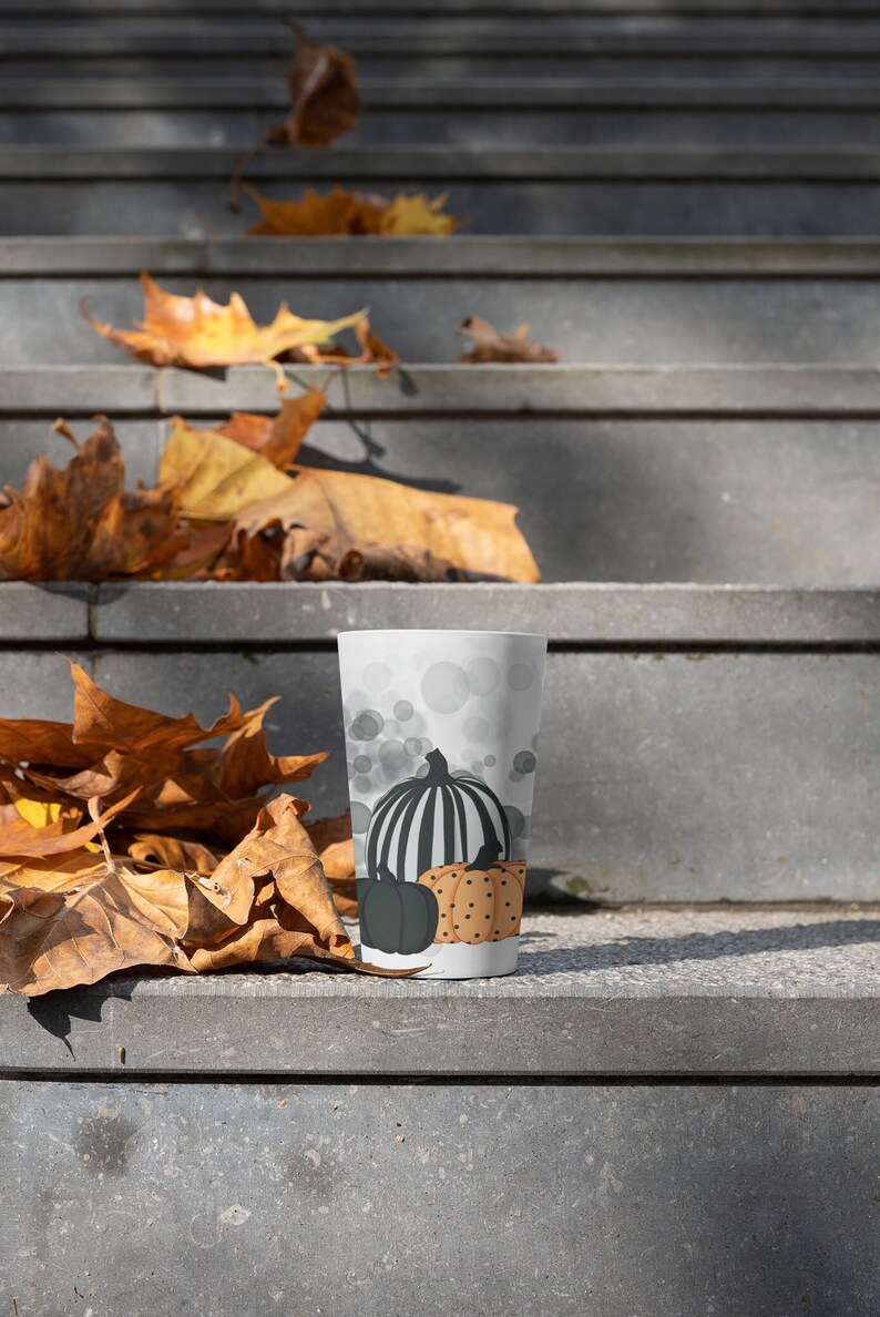 Puede incluir: Un vaso blanco con un dise&ntilde;o de calabaza oto&ntilde;al. El vaso tiene una calabaza a rayas blancas y negras, una calabaza verde oscuro y una calabaza con lunares naranjas. El fondo tiene c&iacute;rculos grises.