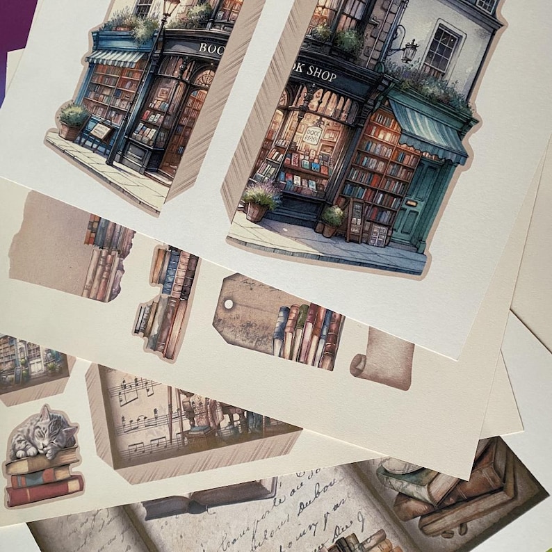 May include: A set of printable paper craft cutouts featuring a whimsical bookstore with a green awning, stacks of books, and a cat curled up on a pile of books. The text "Book Shop" is visible on the storefront.