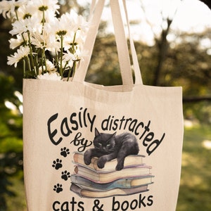 May include: A beige tote bag with a black cat sitting on a stack of books. The bag has the text "Easily distracted by cats & books" printed on it.