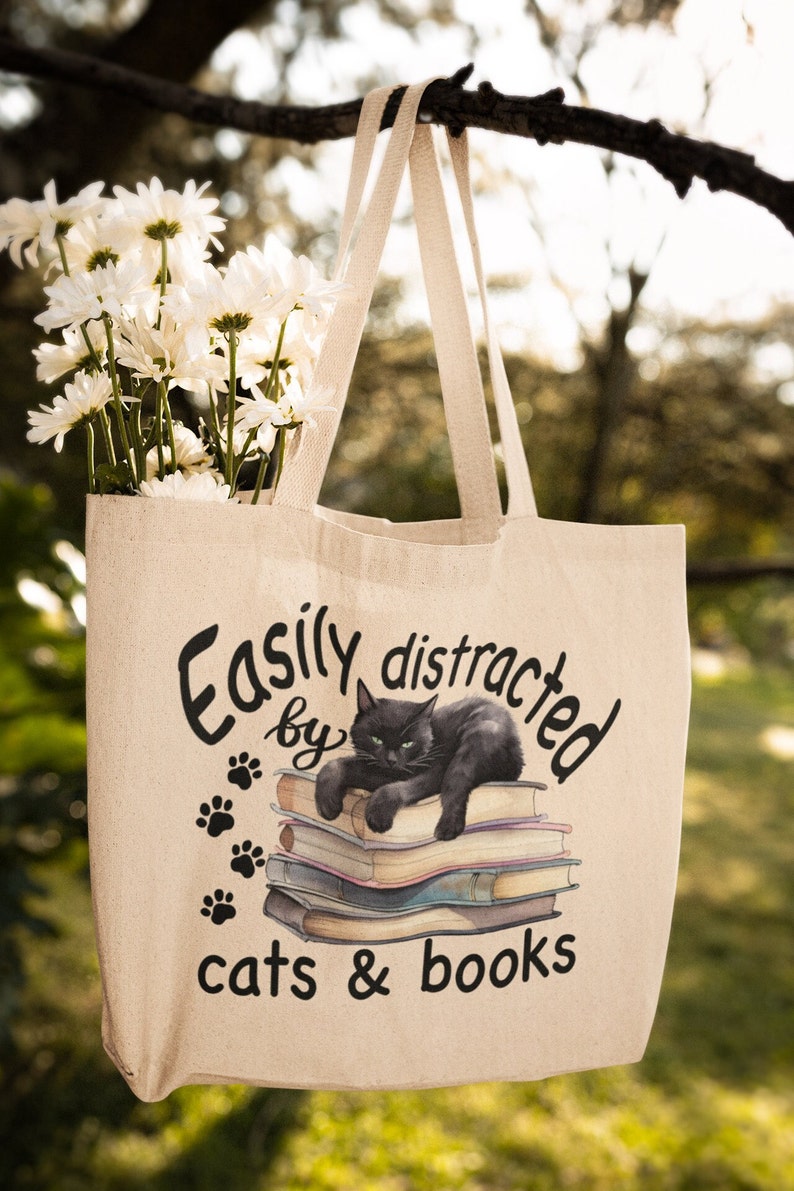May include: A beige tote bag with a black cat sitting on a stack of books. The bag has the text "Easily distracted by cats & books" printed on it.