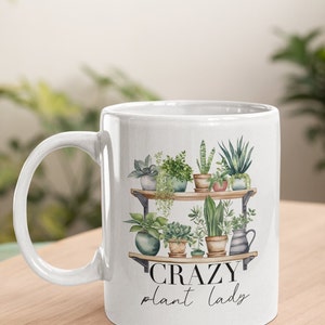 May include: White ceramic mug with a black and white graphic of a shelf with potted plants and the text "CRAZY plant lady".