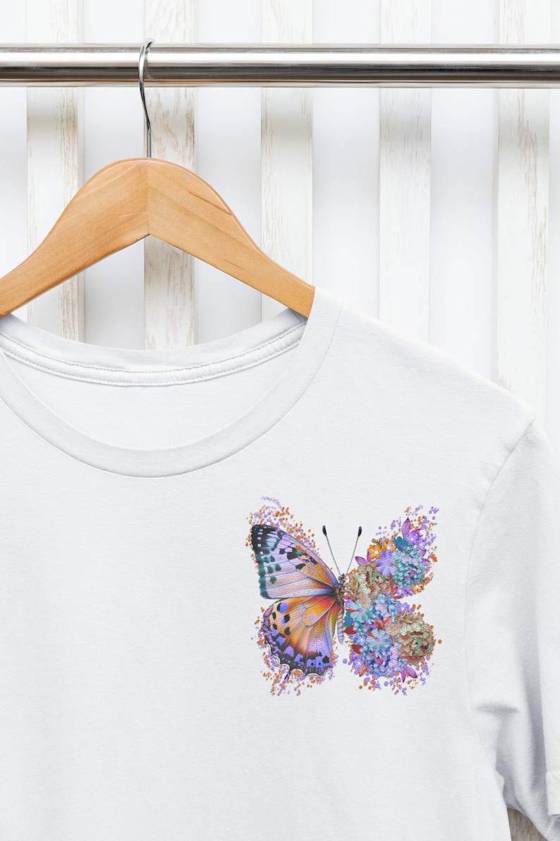 Puede incluir: Camiseta blanca con un gr&aacute;fico de mariposa colorido. La mariposa est&aacute; hecha de flores y tiene acentos morados, naranjas y verdes.