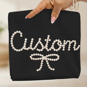 May include: Black tote bag with the word "Custom" spelled out in white pearls, with a pearl bow at the bottom. The bag is held up by a hand with a light skin tone.