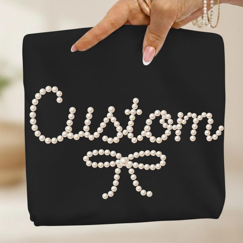May include: Black tote bag with the word "Custom" spelled out in white pearls, with a pearl bow at the bottom. The bag is held up by a hand with a light skin tone.