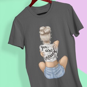 K&ouml;nnte beinhalten: Ein anthrazitfarbenes T-Shirt mit einem Motiv einer Frau mit blonden Haaren in einem Dutt, die Jeans-Shorts und ein wei&szlig;es T-Shirt mit der Aufschrift "you are enough" tr&auml;gt.