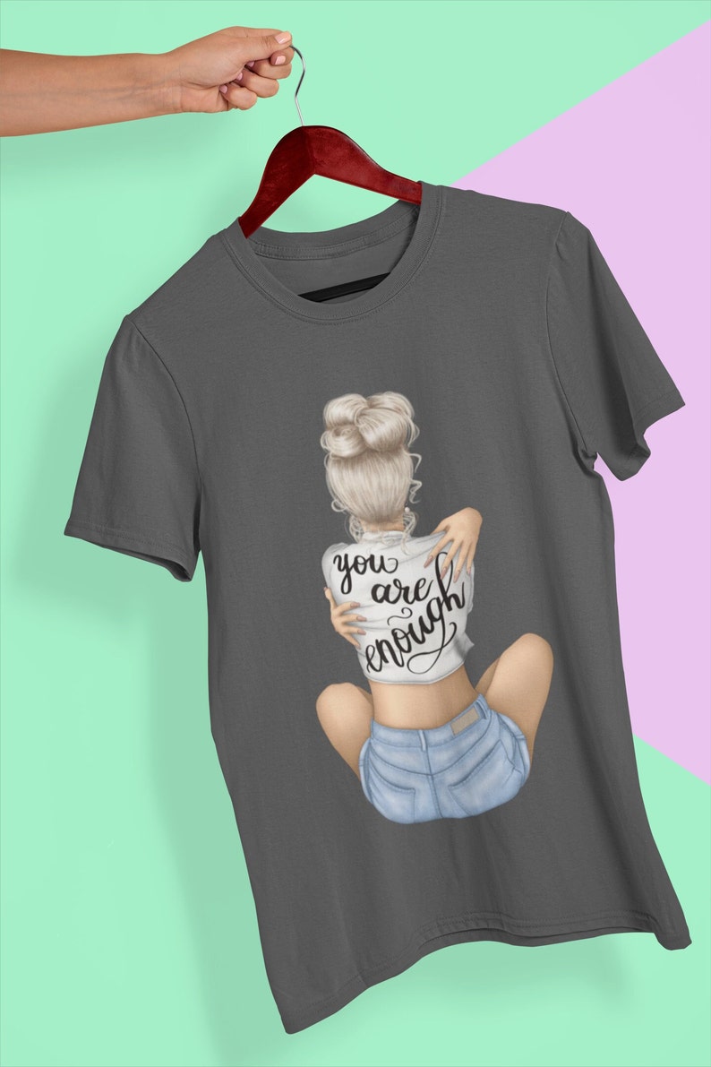 K&ouml;nnte beinhalten: Ein anthrazitfarbenes T-Shirt mit einem Motiv einer Frau mit blonden Haaren in einem Dutt, die Jeans-Shorts und ein wei&szlig;es T-Shirt mit der Aufschrift "you are enough" tr&auml;gt.
