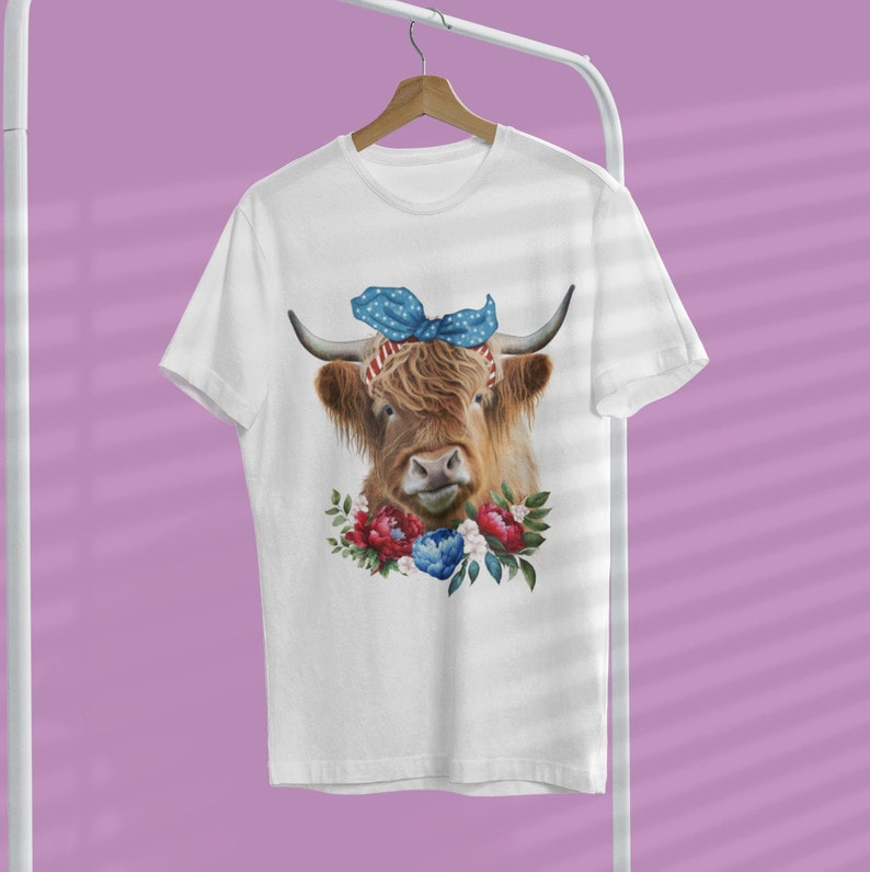 Op de afbeelding: Wit T-shirt met een Schotse hooglander met een blauwe strik en bloemenkrans. De koe heeft bruine vacht en lange hoorns. De krans bevat rode en blauwe bloemen. Het shirt hangt aan een witte hanger.