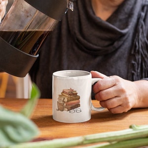 以下が含まれることがあります： 本、メガネ、コーヒーカップのデザインが施された白いセラミックマグ。フレンチプレスからマグカップにコーヒーが注がれています。シーンは木製のテーブルに設定されています。