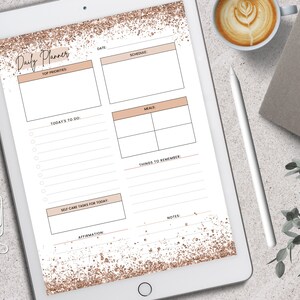 May include: A digital daily planner displayed on a tablet. The planner features sections for top priorities, schedule, meals, and notes, with a rose gold glitter design. A cup of coffee, a pen, and greenery are also visible.