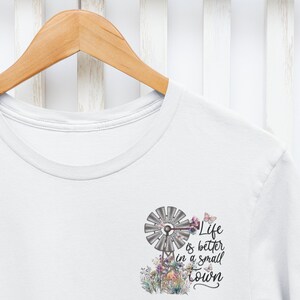 May include: White t-shirt with a graphic of a windmill, flowers, and butterflies. The text on the shirt reads "Life is better in a small town."