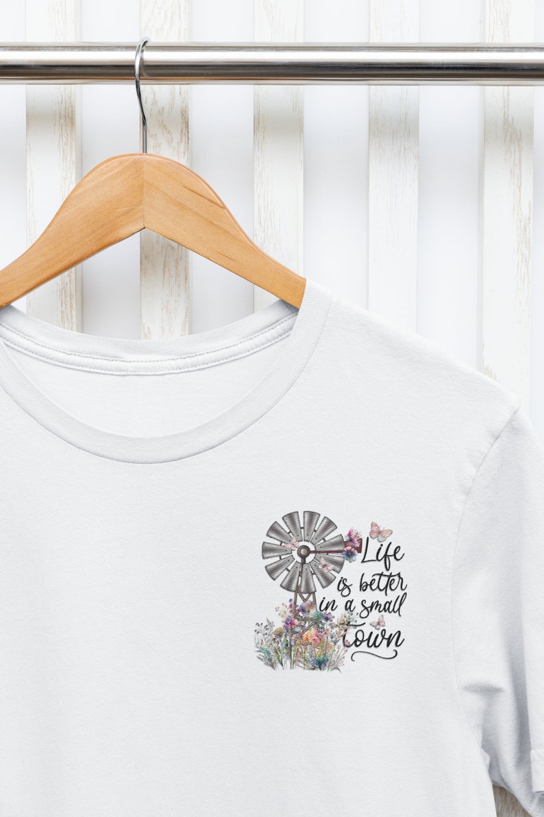May include: White t-shirt with a graphic of a windmill, flowers, and butterflies. The text on the shirt reads "Life is better in a small town."