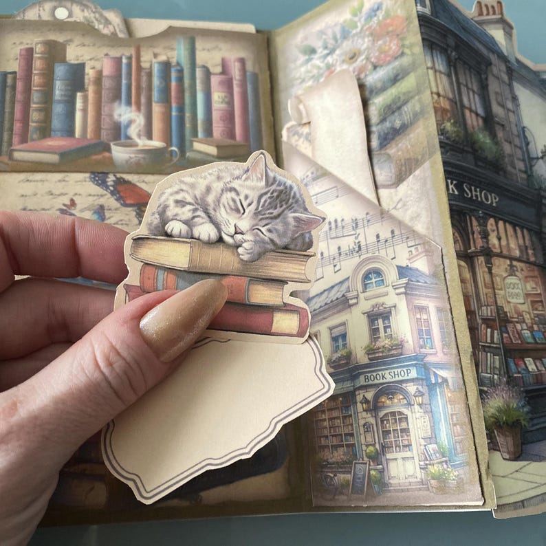 May include: A vintage-style scrapbook with a cat sleeping on a stack of books. The scrapbook features a book shop illustration with the text "Book Shop" and a floral design.