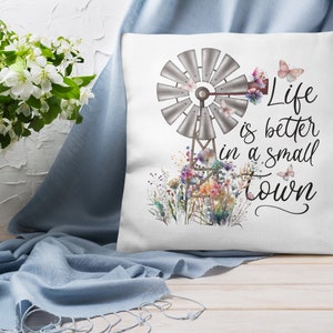 May include: White throw pillow with a watercolor design of a windmill, flowers, and butterflies. The text "Life is better in a small town" is printed on the pillow.