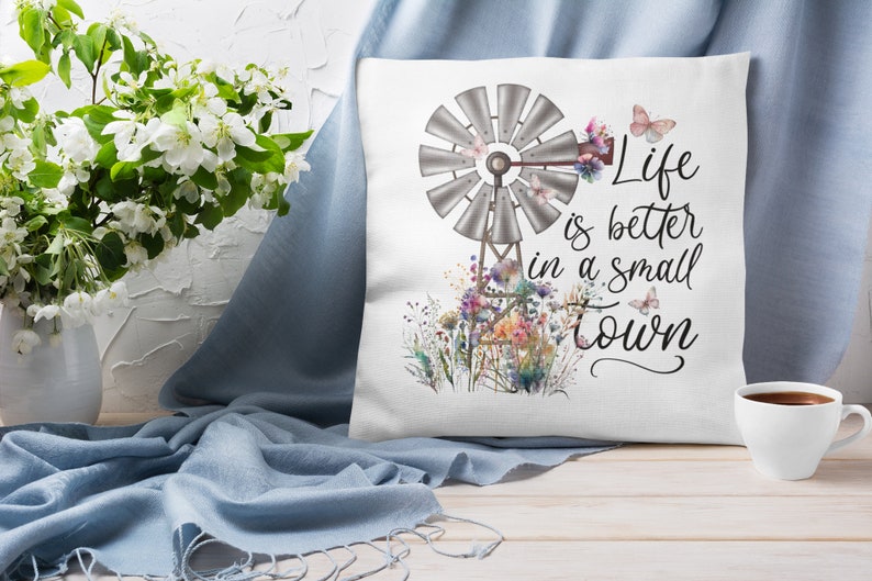 May include: White throw pillow with a watercolor design of a windmill, flowers, and butterflies. The text "Life is better in a small town" is printed on the pillow.