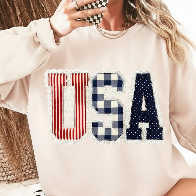 May include: A light pink sweatshirt with the word "USA" appliqued on the front. The "U" is red and white striped, the "S" is blue and white gingham, and the "A" is navy blue with white polka dots. Each letter is outlined with white lace.