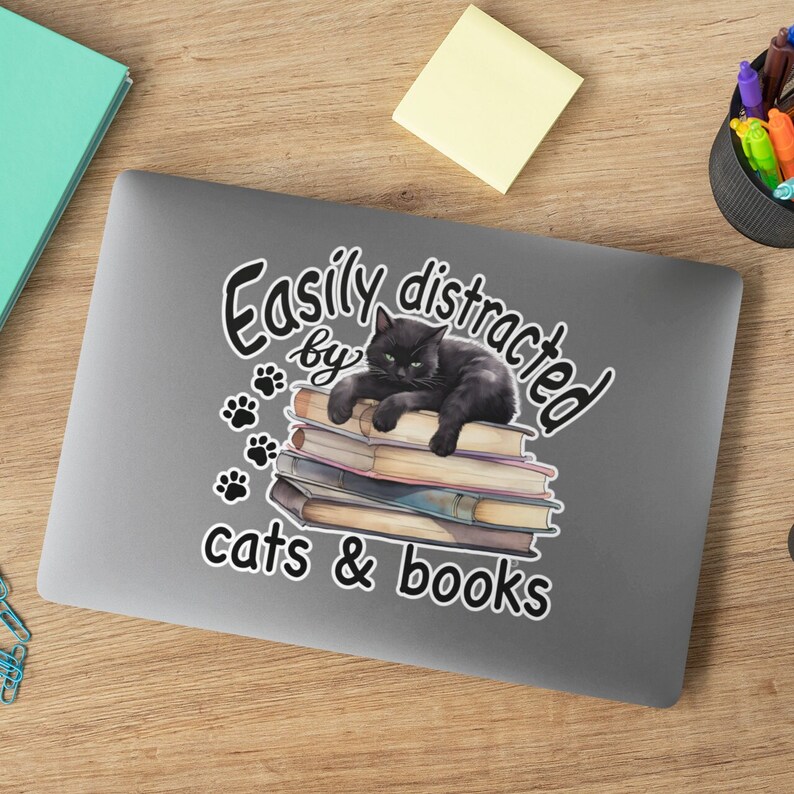May include: A black cat is lying on a stack of books. The text "Easily distracted by cats & books" is written around the cat and books.