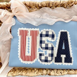 May include: A light blue fabric with the letters "USA" appliqued in red and white stripes, navy blue gingham, and navy blue polka dots. The letters are outlined with white lace trim. The fabric is displayed in a woven basket with a cream-colored ribbon.