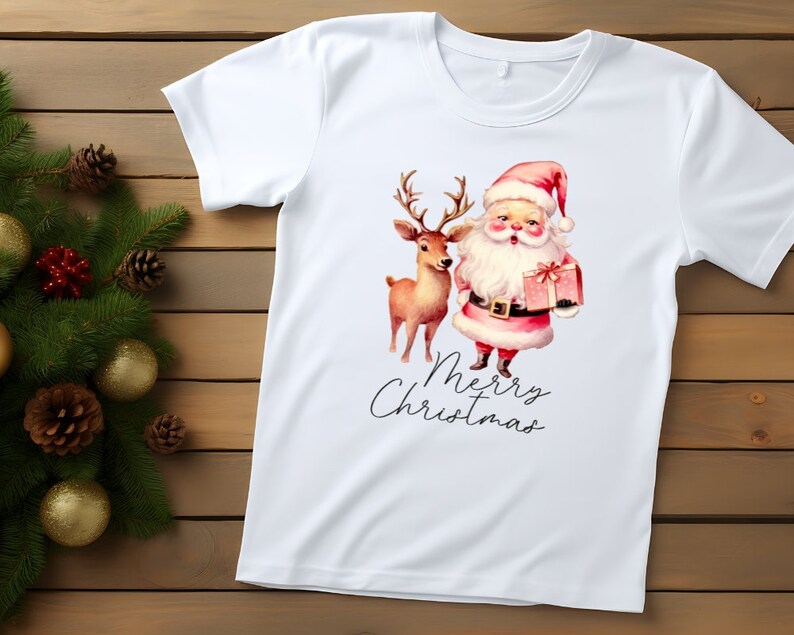 May include: White t-shirt featuring a watercolor illustration of Santa Claus holding a gift box and a reindeer. The words "Merry Christmas" are written in a script font below the image. The shirt is on a wooden surface.
