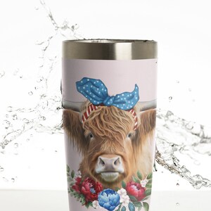 Op de afbeelding: Een lichtroze beker met een Highland-koe met een blauwe strik met sterrenpatroon en een rood-witte gestreepte hoofdband. De koe is omgeven door rode, blauwe en witte bloemaccenten. De beker heeft een zilveren rand.