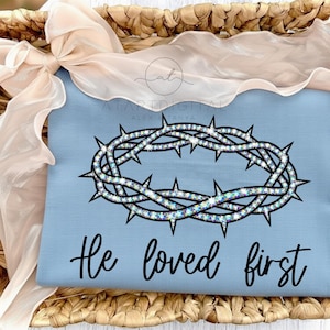 Puede incluir: Un cojín azul claro con un diseño de corona de espinas brillante. El texto "He loved first" está escrito en escritura negra debajo. El cojín está colocado en una cesta de mimbre.
