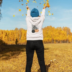 Puede incluir: Una persona con una sudadera blanca con un dise&ntilde;o de calabaza, vaqueros negros y un gorro azul, lanzando hojas amarillas al aire. El fondo muestra un campo de hierba y &aacute;rboles amarillos bajo un cielo azul.