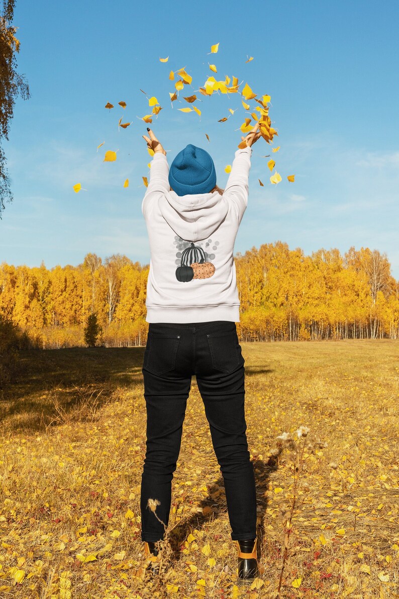 Puede incluir: Una persona con una sudadera blanca con un dise&ntilde;o de calabaza, vaqueros negros y un gorro azul, lanzando hojas amarillas al aire. El fondo muestra un campo de hierba y &aacute;rboles amarillos bajo un cielo azul.