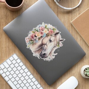 May include: A white horse with a floral crown made of pink, yellow, and blue flowers. The horse's head is turned to the side, and its eyes are closed. The background is a soft, watercolor-like wash of color.