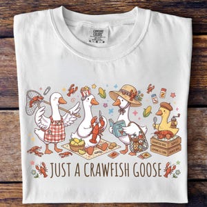 May include: White t-shirt featuring a whimsical illustration of geese preparing a crawfish boil. The design includes geese wearing hats and aprons, holding crawfish, and the text "Just a Crawfish Goose".