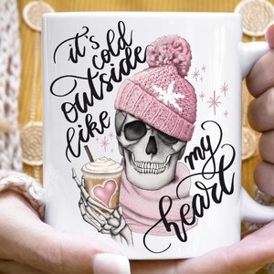 May include: White ceramic mug featuring a skull wearing a pink knit hat and scarf, holding a coffee drink with a heart. The mug has the text "It's cold outside like my heart" in black script.