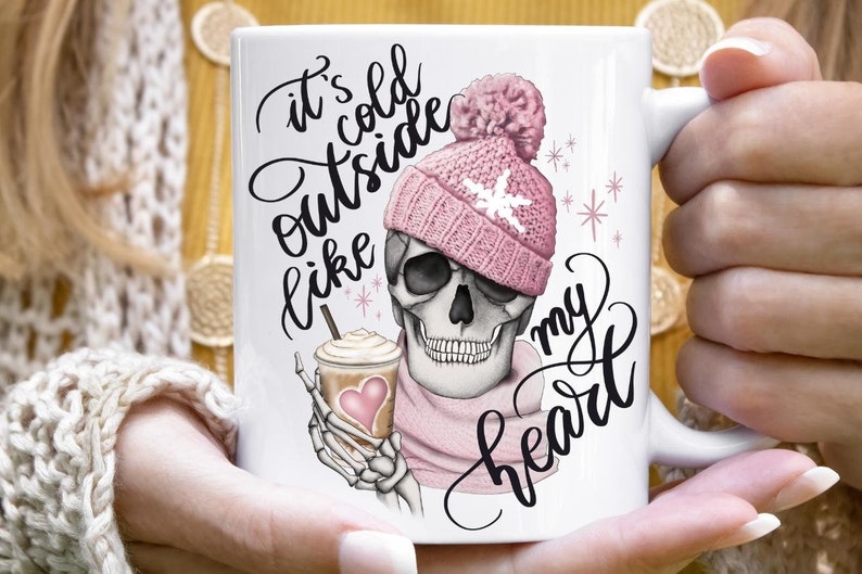 May include: White ceramic mug featuring a skull wearing a pink knit hat and scarf, holding a coffee drink with a heart. The mug has the text "It's cold outside like my heart" in black script.