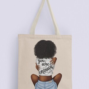 Pu&ograve; includere: Borsa tote in tela beige con manici color crema. La borsa presenta un'illustrazione di una persona con capelli ricci scuri, che indossa una camicia bianca e pantaloncini di jeans. La camicia ha le parole "you are enough" in corsivo.