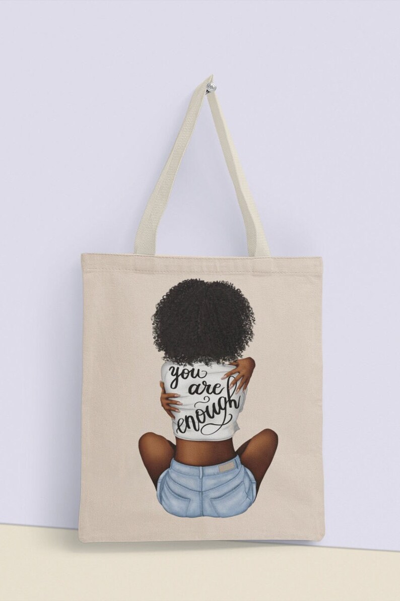 Pu&ograve; includere: Borsa tote in tela beige con manici color crema. La borsa presenta un'illustrazione di una persona con capelli ricci scuri, che indossa una camicia bianca e pantaloncini di jeans. La camicia ha le parole "you are enough" in corsivo.
