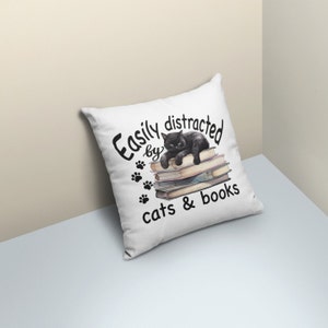 May include: White throw pillow with black text that reads "Easily distracted by cats & books". A black cat is lying on a stack of books.