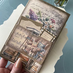 May include: A vintage-style scrapbook with decorative paper accents, including a musical score, a floral design, and a book shop illustration. The scrapbook is open to reveal several pages with different designs.